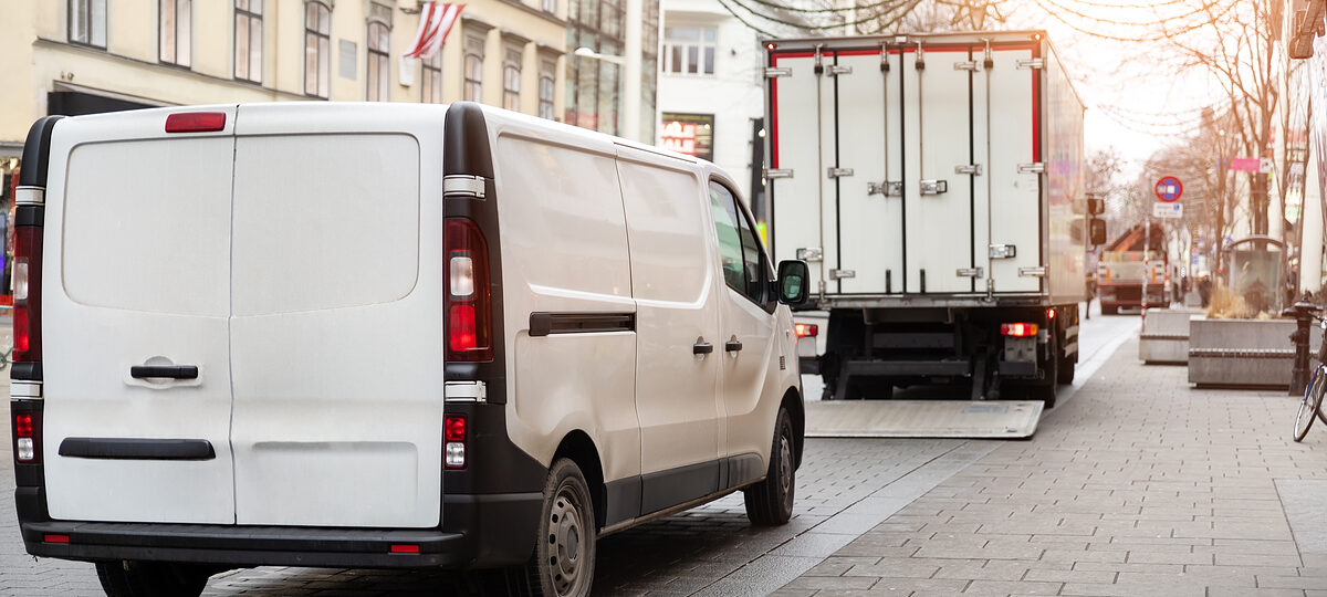 Lieferwagen und Lkw beim Be- oder Entladen auf einer Innenstadtstraße mit Häuserfassaden und Pflastersteinen.