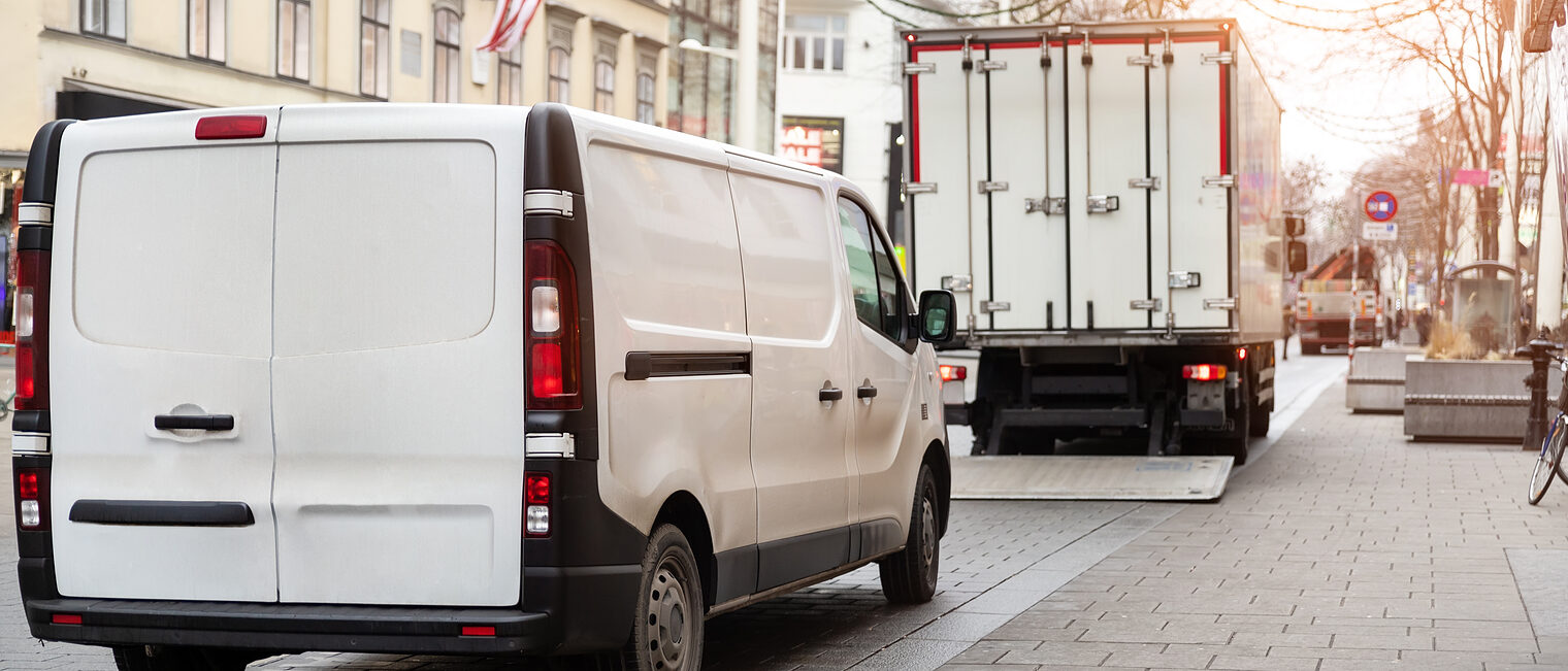 Lieferwagen und Lkw beim Be- oder Entladen auf einer Innenstadtstraße mit Häuserfassaden und Pflastersteinen.