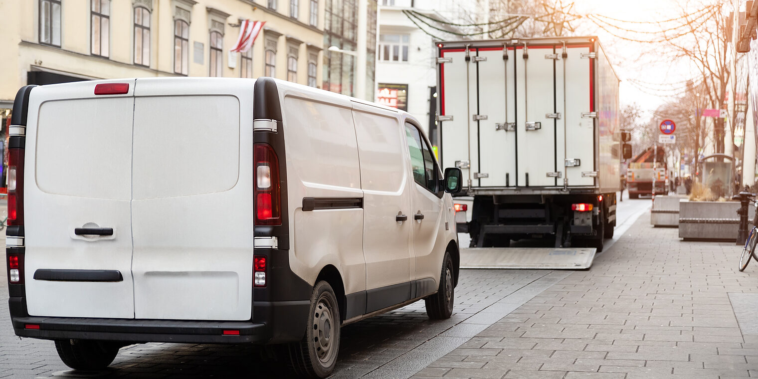 Lieferwagen und Lkw beim Be- oder Entladen auf einer Innenstadtstraße mit Häuserfassaden und Pflastersteinen.