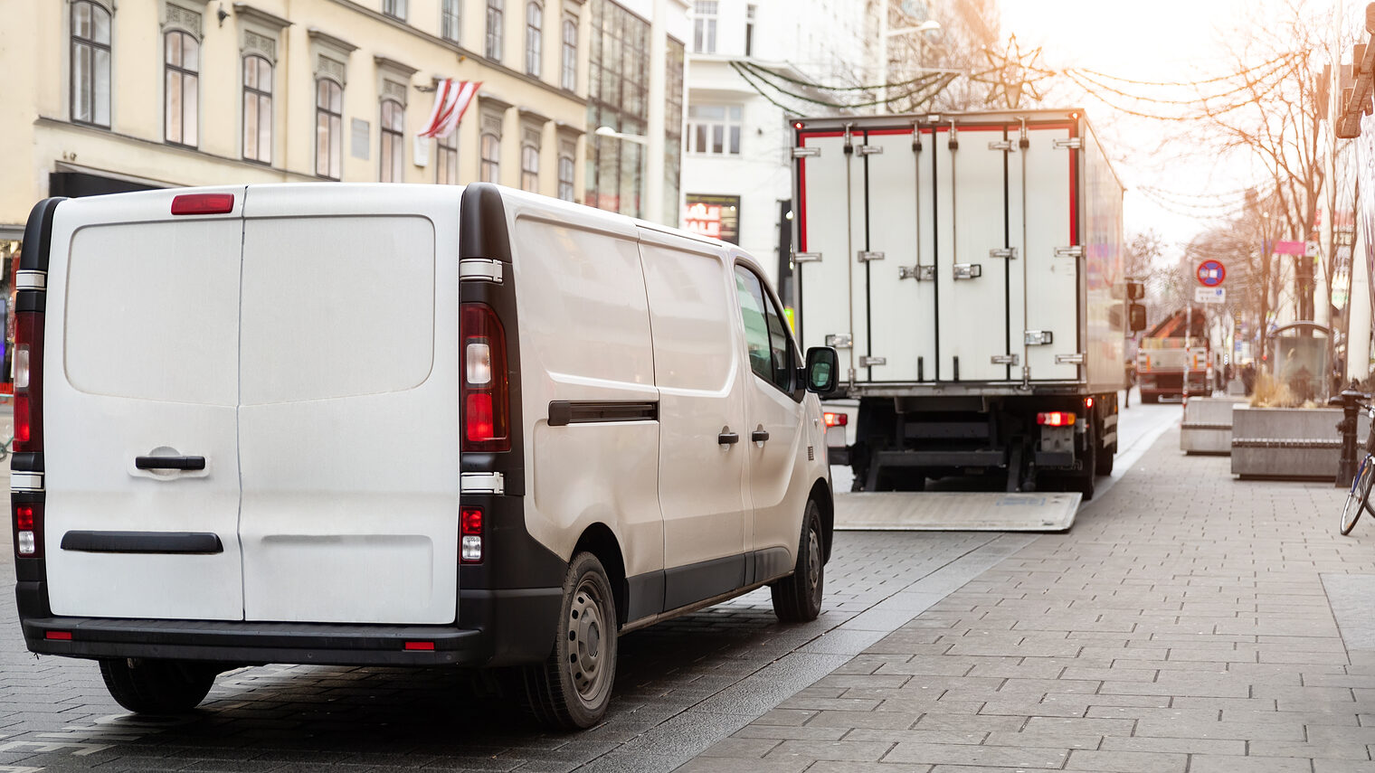 Lieferwagen und Lkw beim Be- oder Entladen auf einer Innenstadtstraße mit Häuserfassaden und Pflastersteinen.