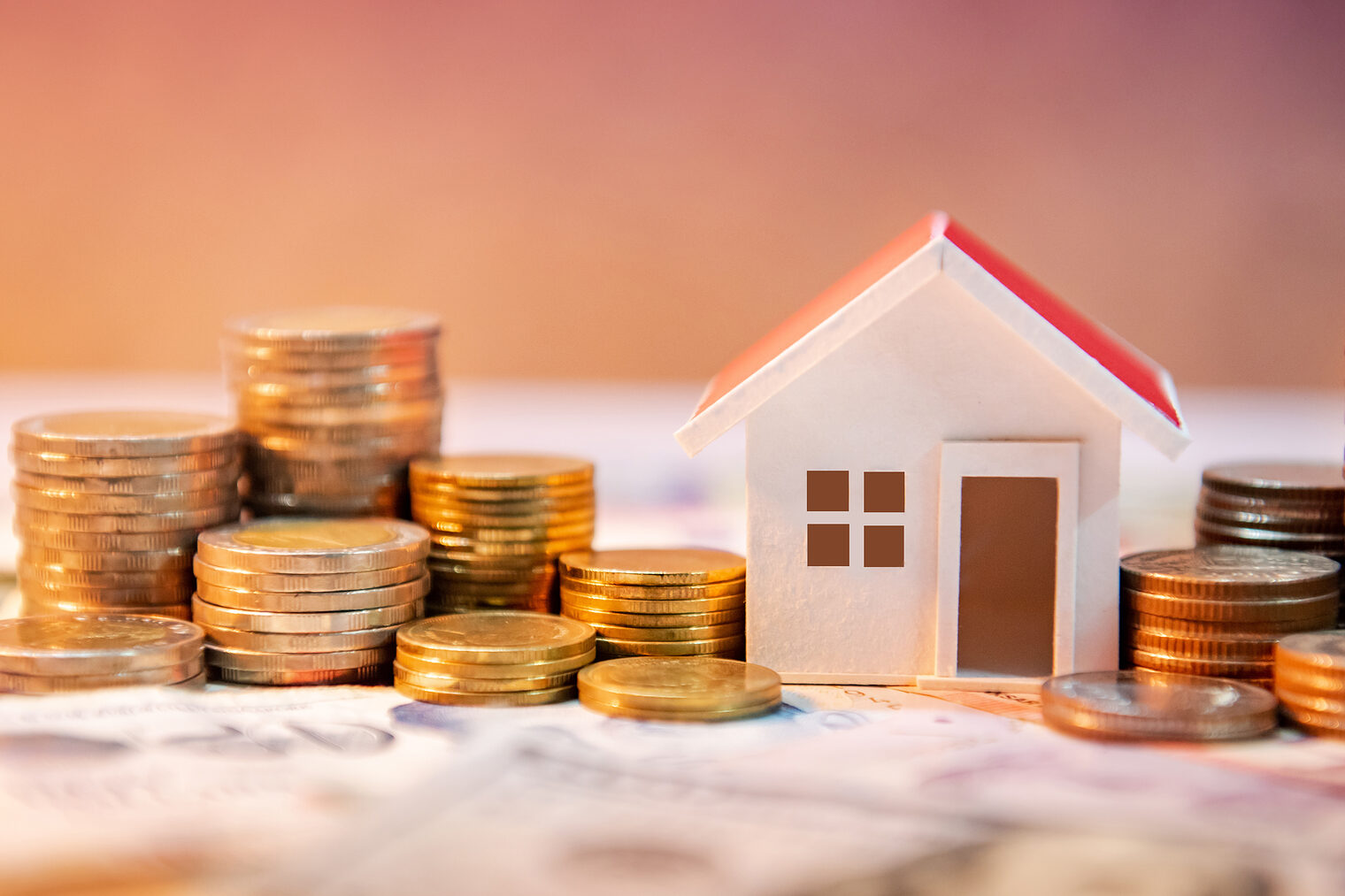 Hausfinanzierung, Investition oder Vermögensaufbau im Fokus Symbolisches Hausmodell zwischen Münzstapeln auf Geldscheinen, Fokus auf Baufinanzierung oder Investitionen