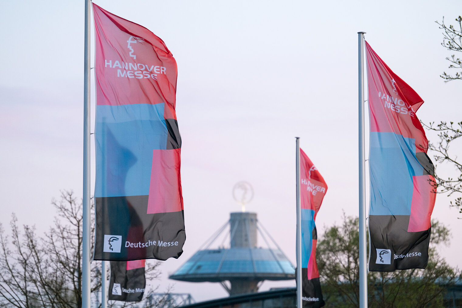 Fahnen der HANNOVER MESSE 2025 wehen vor dem Messezentrum HANNOVER MESSE-Fahnen auf dem Messegelände mit Expo-Dach im Hintergrund bei Abendlicht