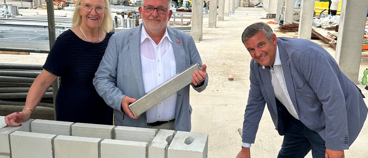 Richtfest Werkstattzentrum III– symbolischer Akt mit Gästen aus dem Handwerk Drei Personen beim symbolischen Mauerbau mit Kelle und Stein auf einer Baustelle mit Rohbau im Hintergrund