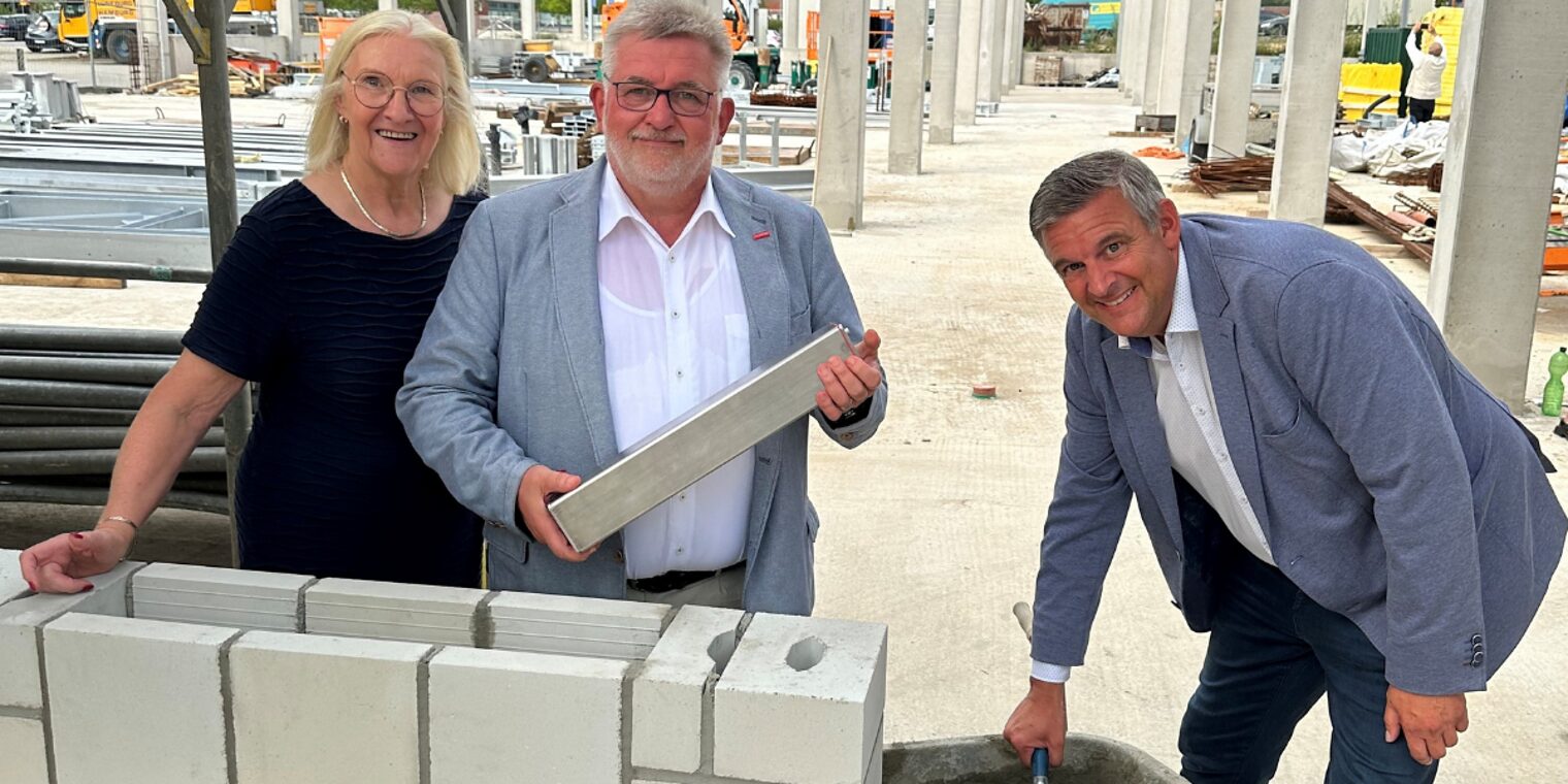 Drei Personen beim symbolischen Mauerbau mit Kelle und Stein auf einer Baustelle mit Rohbau im Hintergrund