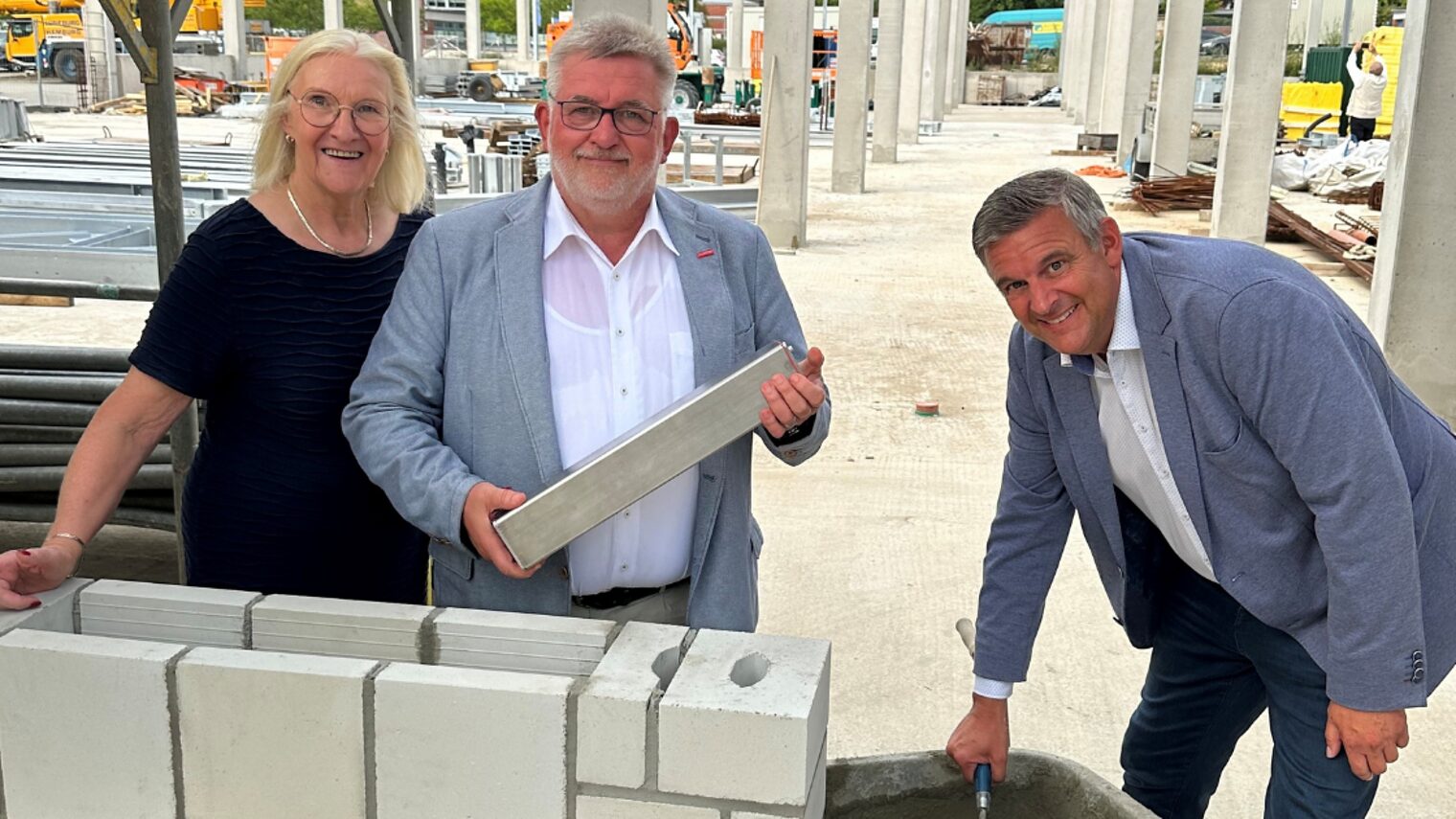 Drei Personen beim symbolischen Mauerbau mit Kelle und Stein auf einer Baustelle mit Rohbau im Hintergrund