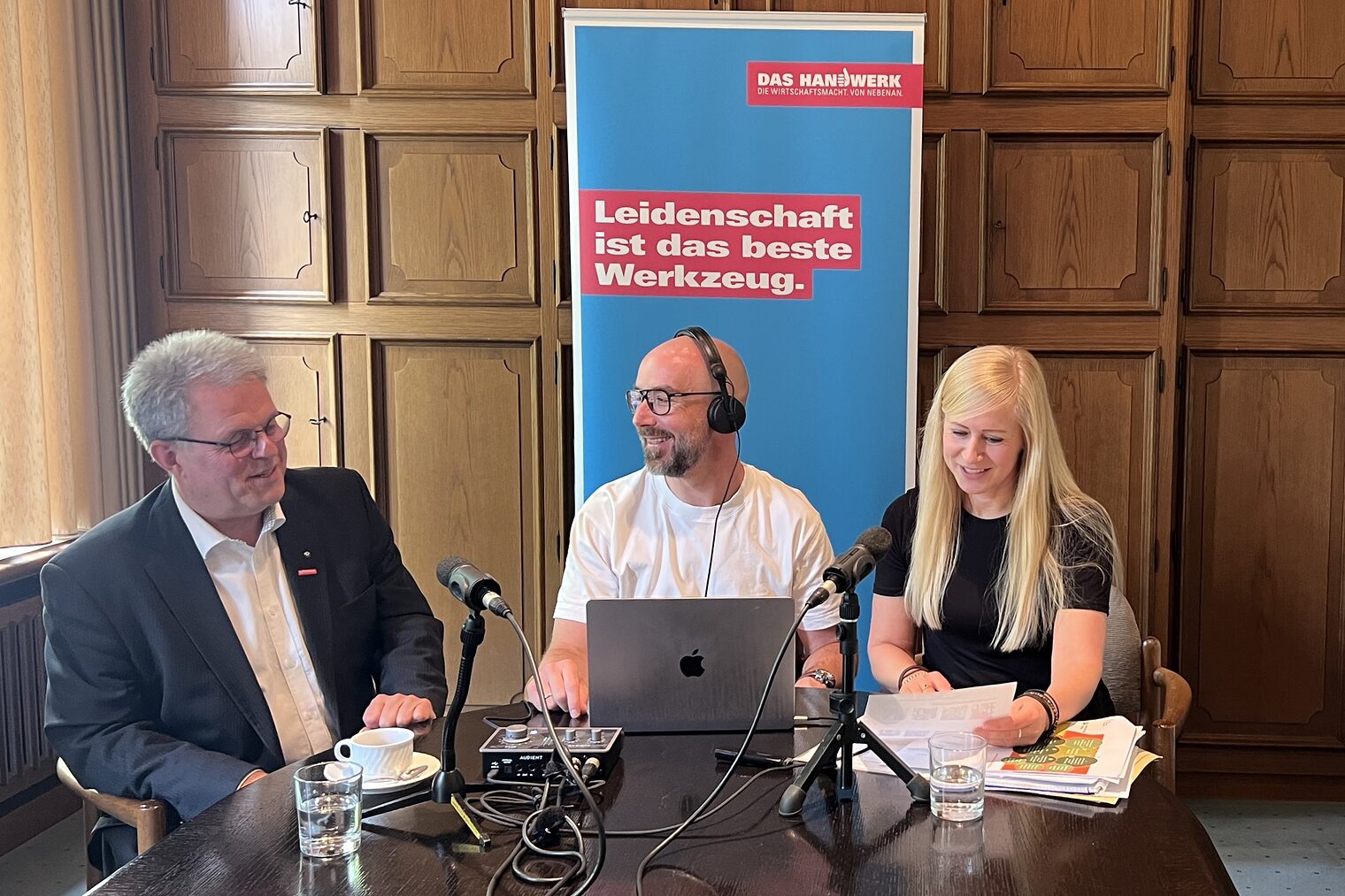 Podcast-Produktion mit Gästen aus dem Handwerk im Interviewformat Drei Personen bei einer Podcast-Aufzeichnung am Tisch mit Mikrofonen, Laptop Roll-Up im Hintergrund.