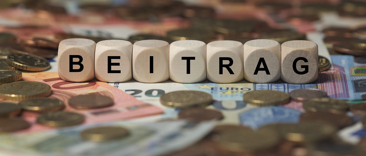 Holzwürfel mit dem Wort „Beitrag“ auf Euro-Banknoten und Münzen als Symbol für Beiträge und Finanzierung.