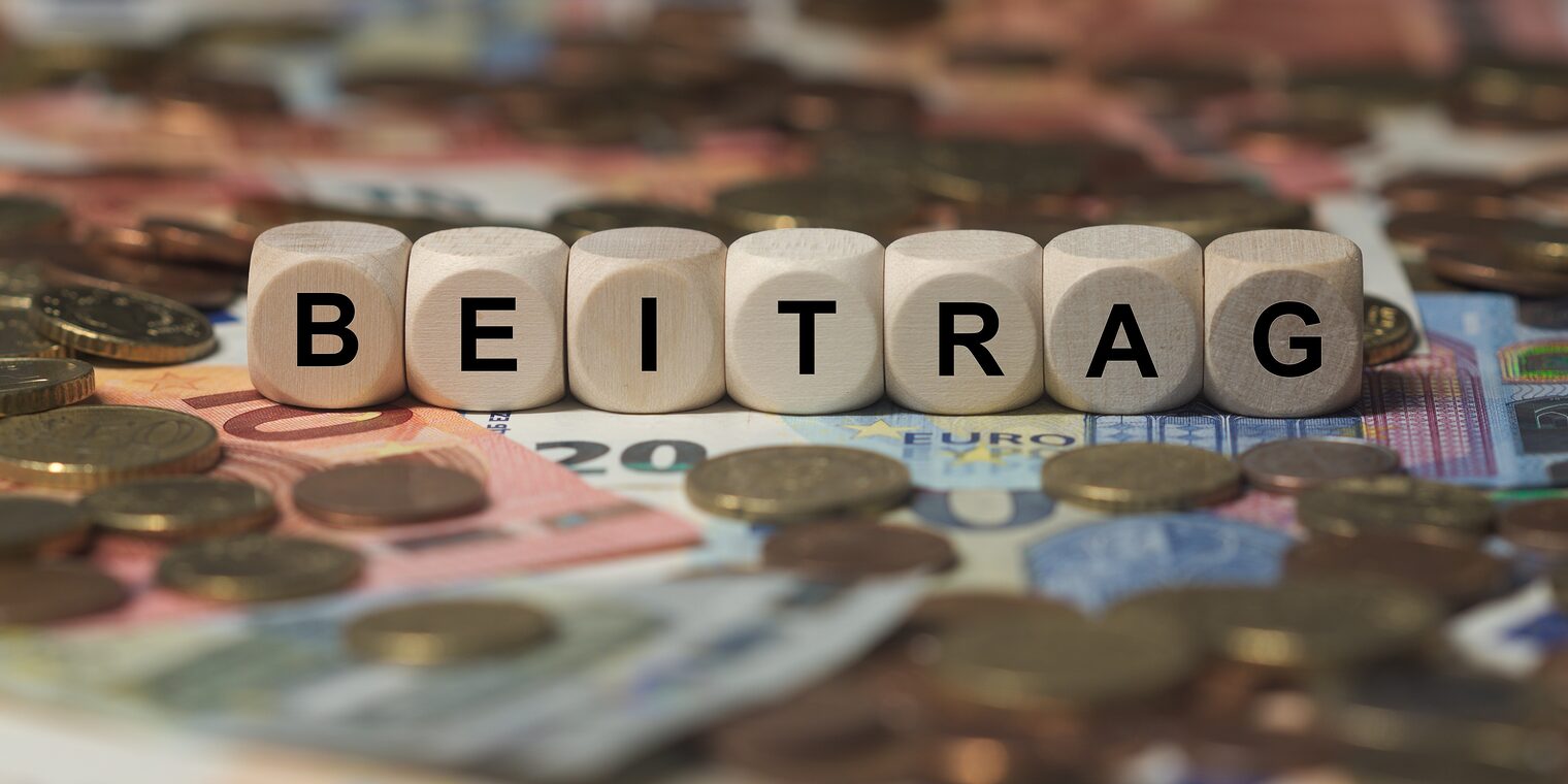 Holzwürfel mit dem Wort „Beitrag“ auf Euro-Banknoten und Münzen als Symbol für Beiträge und Finanzierung.