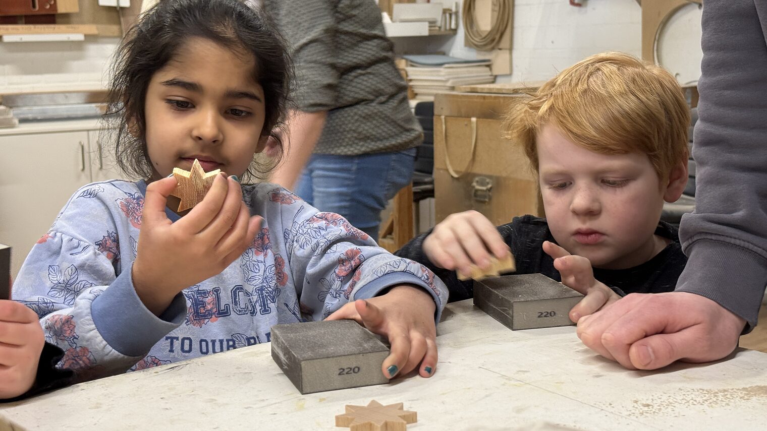 Zwei Kindergartenkinder blicken in einer Werkstatt stolz auf ihre selbst gemachten Holzsterne.