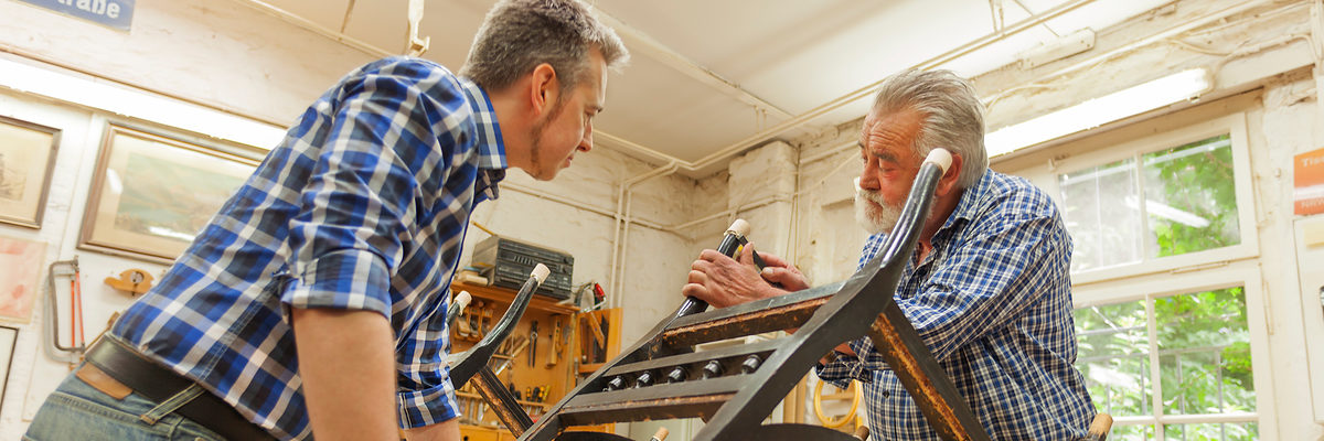 Älterer Handwerker zeigt jüngerem Handwerker eine Handwerkstechnik an einem Stuhl.